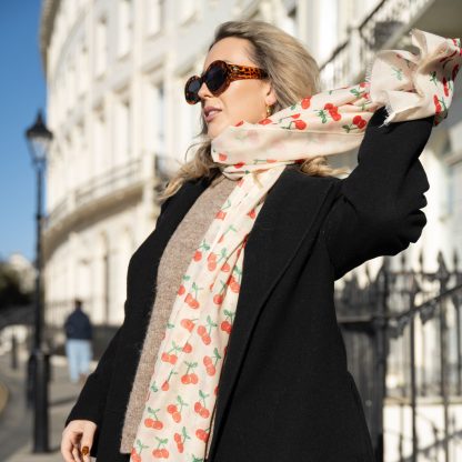 Blonde lady standing outside white buildings in chelsea, wearing an all back outfit, taupe jumper and a hand printed cherries design scarf with black sunglasses on