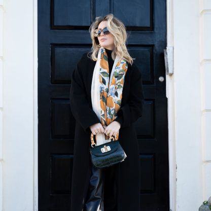 Blonde lady standing outside white buildings in chelsea, wearing an all back outfit, taupe jumper and a hand printed lemons and hydrangeas design scarf with black sunglasses on