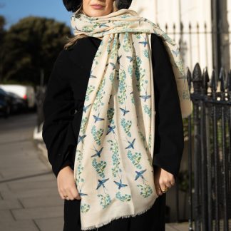 Blonde lady standing outside white buildings in chelsea, wearing an all back outfit, taupe jumper and a hand printed blue birds design scarf with black sunglasses on