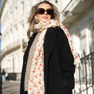 Blonde lady standing outside white buildings in chelsea, wearing an all back outfit, taupe jumper and a hand printed cherries design scarf with black sunglasses on