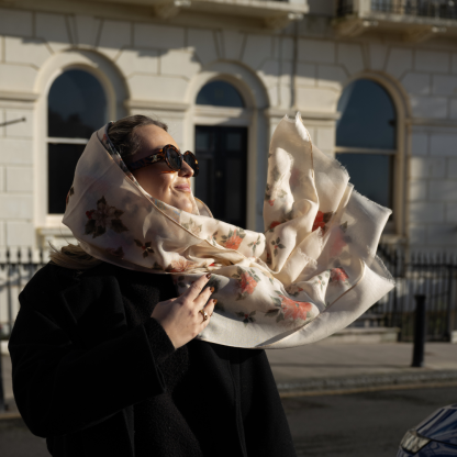 Blonde lady standing outside white buildings in chelsea, wearing an all back outfit, taupe jumper and a hand printed poinsettia design scarf with black sunglasses on