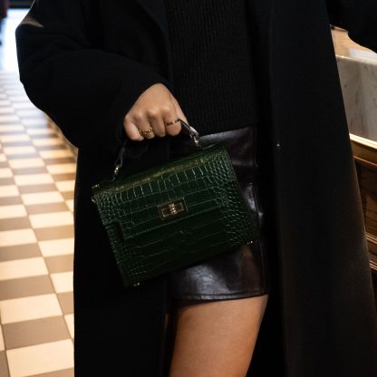 Blonde lady holding an emerald green genuine italian leather croc crossbody bag, standing at a bar in chelsea in black outfit , holding a glass of champagne