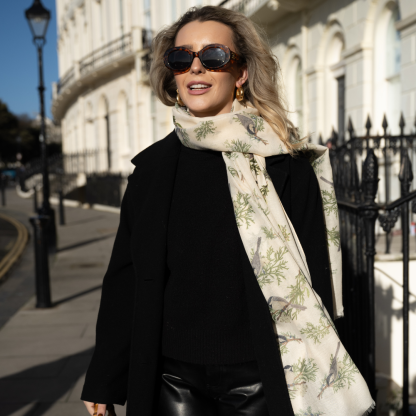 Blonde lady standing outside white buildings in chelsea, wearing an all back outfit, taupe jumper and a hand printed finch birds design scarf with black sunglasses on