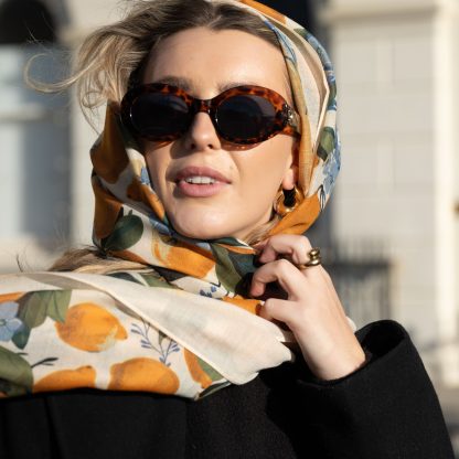 Blonde lady standing outside white buildings in chelsea, wearing an all back outfit, taupe jumper and a hand printed lemons and hydrangeas design scarf with black sunglasses on