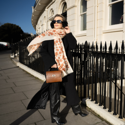 Blonde lady standing outside white buildings in chelsea, wearing an all back outfit, taupe jumper and a hand printed gingerbread man design scarf with black sunglasses on holing a tan italian leather crossbody bag