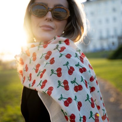 Blonde lady standing outside white buildings in chelsea, wearing an all back outfit, taupe jumper and a hand printed cherries design scarf with black sunglasses on