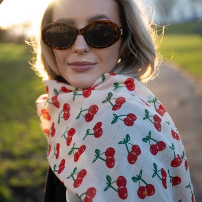 Blonde lady standing outside white buildings in chelsea, wearing an all back outfit, taupe jumper and a hand printed cherries design scarf with black sunglasses on