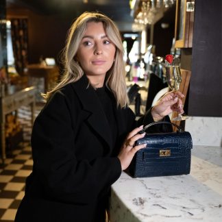 Blonde lady holding navy blue genuine italian leather croc crossbody bag, standing at a bar in chelsea in black outfit , holding a glass of champagne