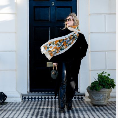 Blonde lady standing outside white buildings in chelsea, wearing an all back outfit, taupe jumper and a hand printed lemons and hydrangeas design scarf with black sunglasses on