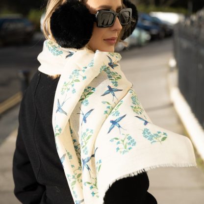 Blonde lady standing outside white buildings in chelsea, wearing an all back outfit, taupe jumper and a hand printed blue birds design scarf with black sunglasses on