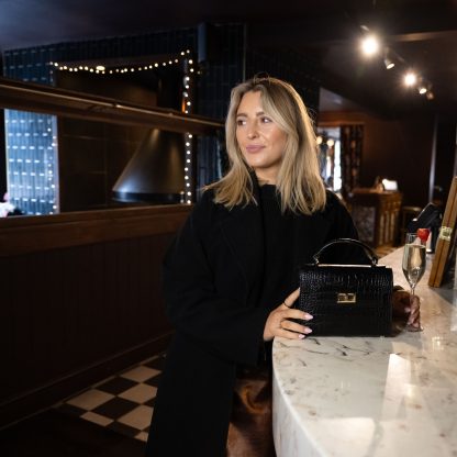 Blonde lady holding a black genuine italian leather croc crossbody bag, standing at a bar in chelsea in black outfit , holding a glass of champagne