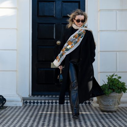 Blonde lady standing outside white buildings in chelsea, wearing an all back outfit, taupe jumper and a hand printed lemons and hydrangeas design scarf with black sunglasses on