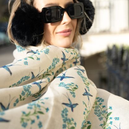 Blonde lady standing outside white buildings in chelsea, wearing an all back outfit, taupe jumper and a hand printed blue birds design scarf with black sunglasses on