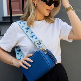 Blonde lady wearing black trousers, white t shirt, grey jumper, black sunglasses with a royal blue italian leather bag and a Blue butterflies bag strap